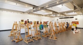 Spacious art classroom with rows of wooden easels, bright overhead lighting, student artists sketching and painting, and colorful artwork displayed on white walls