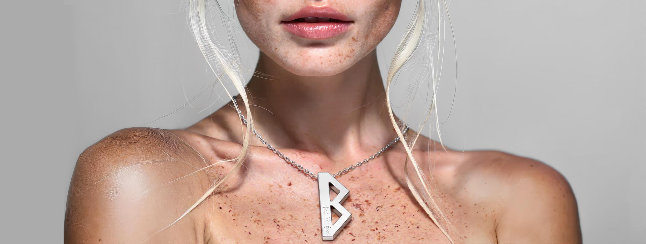 Photorealistic 3D lifestyle rendering of blonde woman with freckles wearing silver necklace with large stylized “B” pendant – close-up shot