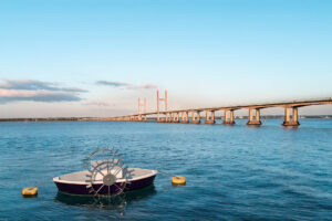 Photorealistic 3D rendering of modern purple and white conceptual paddlewheel boat with large metal paddle wheel, yellow anchor buoys and massive cable-stayed bridge in the background on calm water