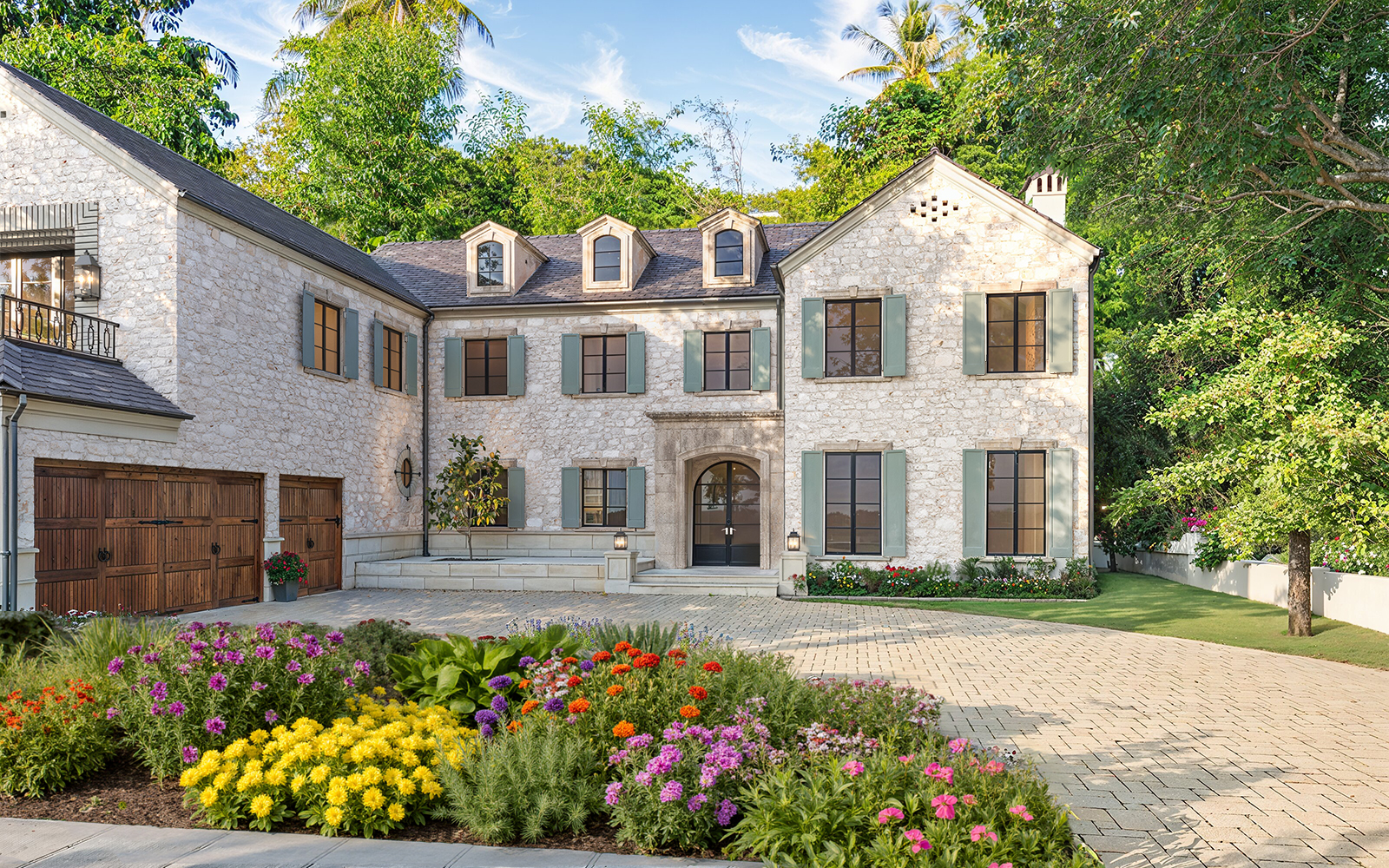 Photorealistic 3D rendering of luxurious two-story stone Raddon Residence Option A with light beige stone facade, sage green shutters, dark slate roof, wooden garage doors, elegant brick driveway and vibrant colorful flower beds in full bloom