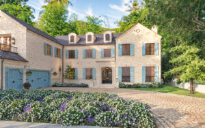 Photorealistic 3D rendering of luxurious two-story stone Raddon Residence Option A with light beige stone facade, blue shutters, dark slate roof, elegant arched entrance, cobblestone driveway and vibrant colorful flower beds in full bloom