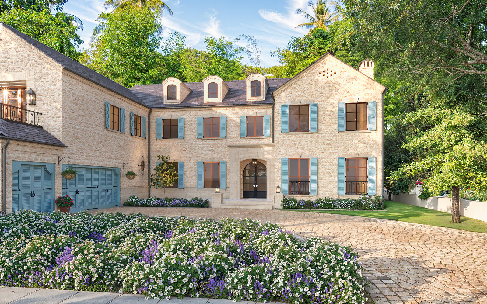 Photorealistic 3D rendering of luxurious two-story stone Raddon Residence Option A with light beige stone facade, blue shutters, dark slate roof, elegant arched entrance, cobblestone driveway and vibrant colorful flower beds in full bloom