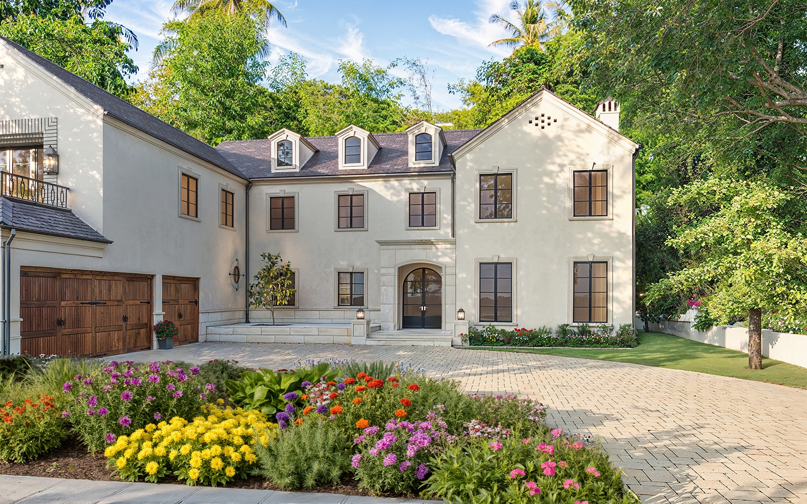 Photorealistic 3D rendering of luxurious two-story cream Raddon Residence Option B with dark slate roof, dormer windows, wooden garage doors, elegant landscaping and vibrant colorful flower beds