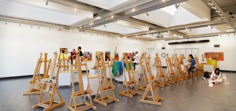 Spacious art classroom with rows of wooden easels, bright overhead lighting, student artists sketching and painting, and colorful artwork displayed on white walls