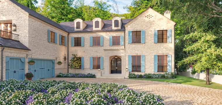 Photorealistic 3D rendering of luxurious two-story stone Raddon Residence Option A with light beige stone facade, blue shutters, dark slate roof, elegant arched entrance, cobblestone driveway and vibrant colorful flower beds in full bloom