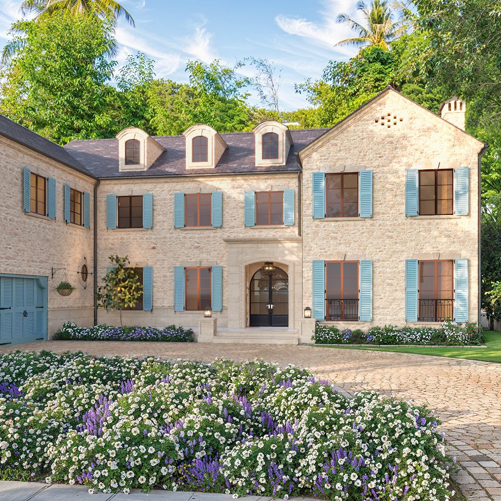 Photorealistic 3D rendering of luxurious two-story stone Raddon Residence Option A with light beige stone facade, blue shutters, dark slate roof, elegant arched entrance, cobblestone driveway and vibrant colorful flower beds in full bloom